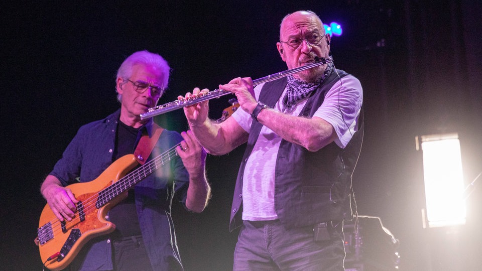 Rechts im Bild spielt ein Mann eine Querflöte, links hinter ihm steht ein Mann mit einer Gitarre